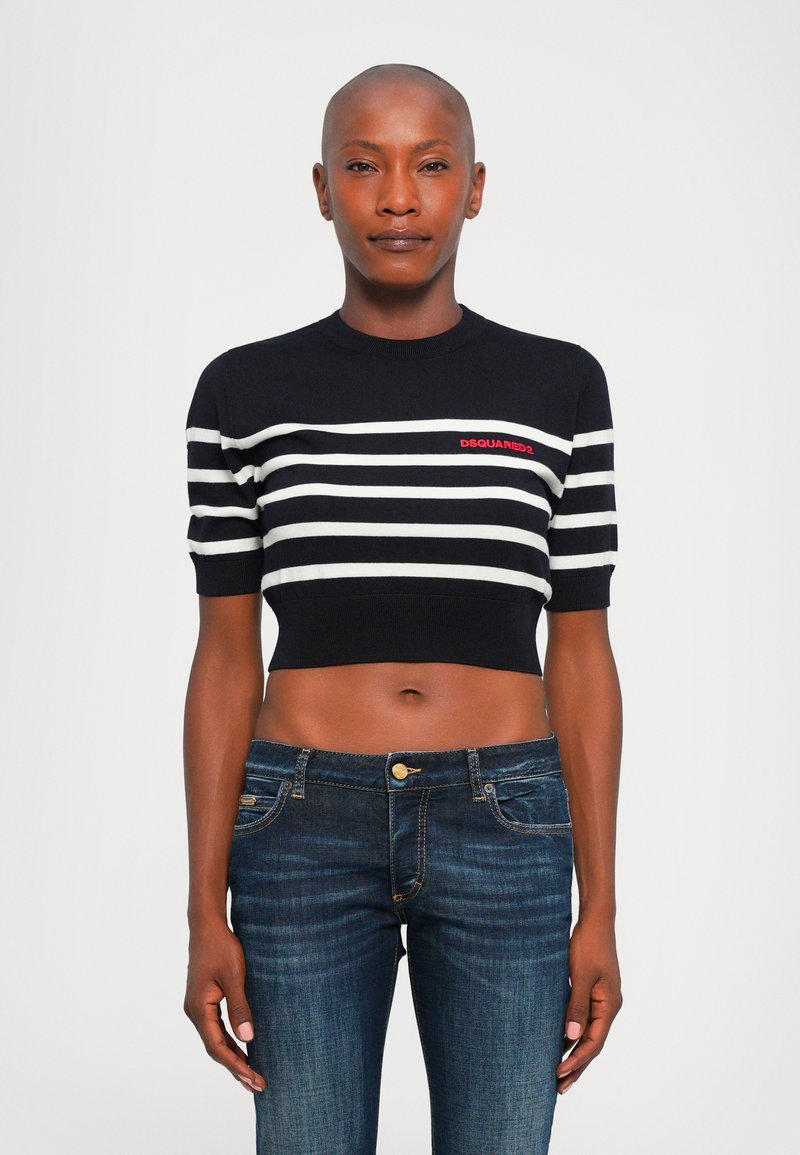 Woman with shaved head wearing a black and white striped cropped sweater and dark blue jeans, standing against a plain background.