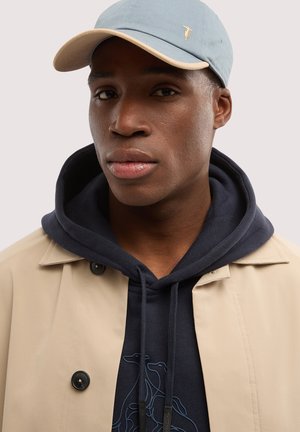 Młody mężczyzna w jasnoniebieskiej czapce, granatowej bluzie z kapturem z motywem psa w stylu linii oraz beżowym trenczu na jednolitym tle.