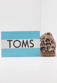 Carte de marque TOMS bleu et blanc à côté d’un talon de chaussure slip-on à imprimé léopard sur une surface et un fond blancs.