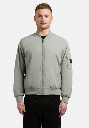 Mężczyzna noszący jasnoszarą kurtkę bomber z przednim zamkiem błyskawicznym i zamykaną kieszenią na lewym rękawie, w czarnych spodniach, stojący na białym tle.