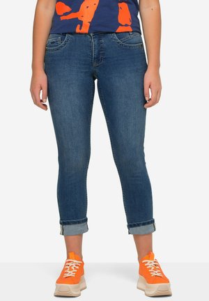 Tmavě modré džíny s cropped fit, s ohrnutými manžetami a jemným vyblednutím, v kombinaci s jasně oranžovými teniskami.