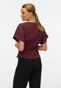 Burgundijas blūze ar īsām, platajām piedurknēm un pieguļošu dizainu, izgatavota no gludas auduma. Valkāta ar melniem biksēm, parādot tīras šūšanas detaļas.