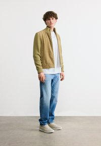 Νέος άνδρας που στέκεται, φοράει μπεζ ζακέτα με φερμουάρ και πλεκτά μανίκια, λευκό T-shirt, ξεβαμμένο μπλε τζιν και μπεζ αθλητικά παπούτσια, σε λευκό φόντο.