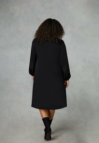 Černé šaty s dlouhými rukávy, které mají krajkové akcenty. Rovný střih, délka ke kolenům a hladká textura. Nosené s černou obuví.