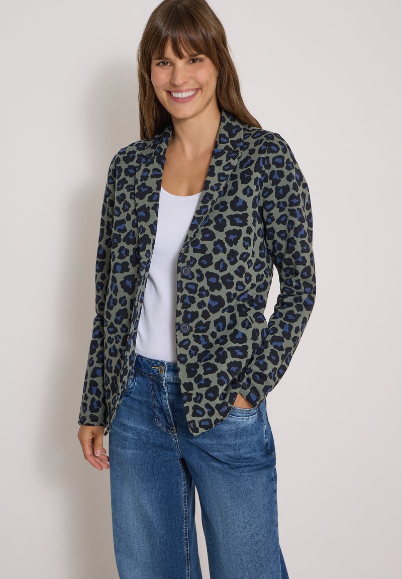 Femme souriante portant un blazer bleu et noir à imprimé léopard sur un haut blanc et un jean bleu, debout avec une main dans sa poche.