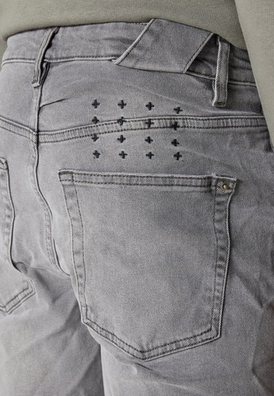 Pantaloni in denim grigio con design della tasca posteriore caratterizzato da motivi incrociati neri. I dettagli includono cuciture standard e chiusura con bottone in metallo.