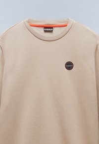 Sudadera beige hecha de una tela suave. Tiene un cuello redondo, una textura sutil y un logotipo circular en colores contrastantes en la parte frontal.