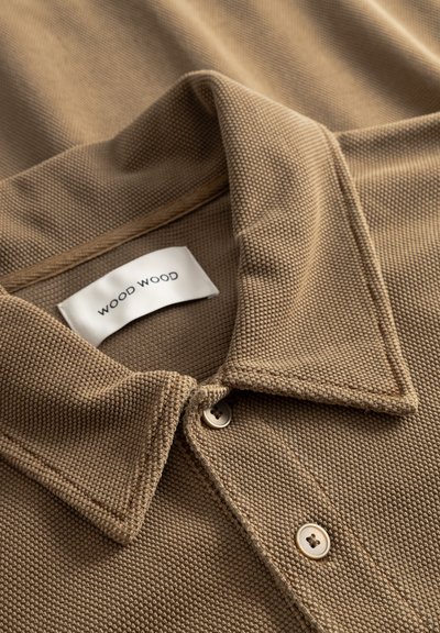 Beige pamut póló texturált szövettel, klasszikus gallérral, két gombbal és egyszerű betűtípusú "WOOD WOOD" címkével.