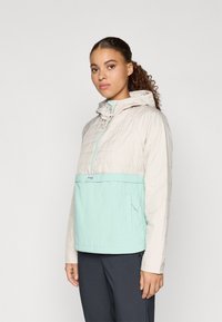 Lahka dvo tona jakna v kremni in mentolno zeleni barvi, opremljena s kapuco, zapiranjem na poln zadrgo, prešitimi teksturami in sprednjim žepom.