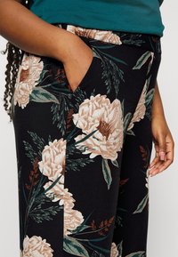 Pantalons à motif floral noir avec de grandes pivoine crèmes et des feuilles vertes. Comprend une poche latérale et un tissu doux et extensible.