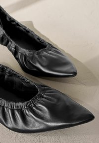 Schwarze Lederballerinas mit spitzem Zehenbereich, gerafftem elastischen Rand und glatter Oberfläche, auf einer hellen Oberfläche platziert.
