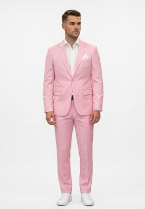 Hombre de pie vistiendo un traje rosa claro con una camisa blanca de vestir, un pañuelo blanco en el bolsillo y zapatillas blancas, sobre un fondo blanco liso.