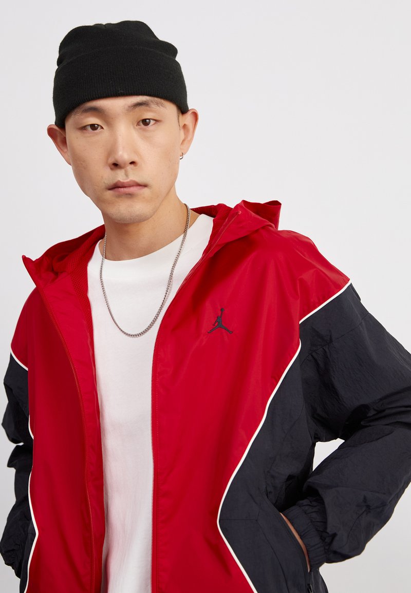 Giacca a vento rossa e nera con zip frontale, cappuccio e logo sul petto. Presenta piping bianco e polsini elasticizzati. Indossata sopra una t-shirt bianca.