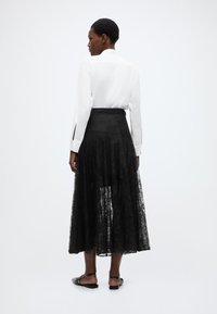 Μαύρη φούστα από δαντέλα, μέχρι τον αστράγαλο με ροή σχεδίου· συνδυασμένη με λευκό πουκάμισο μακρυμάνικο με λεπτομέρεια κοψίματος στα μανίκια.