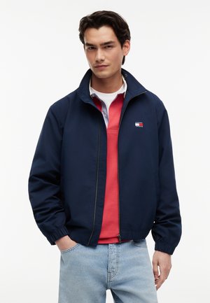 Fiatal férfi, tengerészkék cipzáras dzsekit visel piros ing felett és világoskék farmert, egyik kezét a zsebében tartva fehér háttér előtt áll.