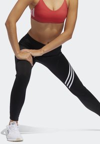 Nainen venyttelee jalkojaan, päällä punainen urheiluhaalari, mustat leggingit valkoisilla raitoineen ja valkoiset urheilukengät yksinkertaisella taustalla.