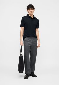 Jeune homme portant un polo noir et un pantalon gris, tenant un sac cabas noir, debout devant un fond uni clair.