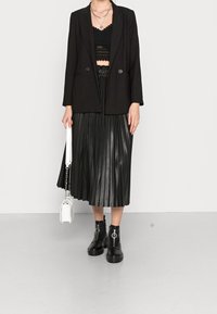 Osoba w czarnej koronkowej bluzce, czarnym żakiecie, czarnej plisowanej spódnicy midi, czarnych botkach za kostkę i trzymająca białą torebkę.