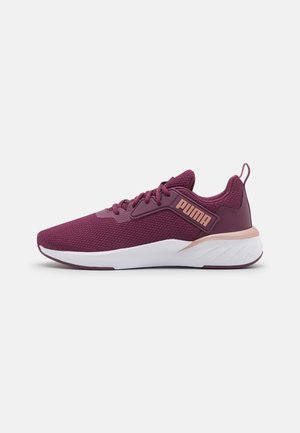 Zapatilla de correr Puma morada con parte superior de malla, cierre de cordones, suela blanca y etiqueta con logo rosa en el lateral sobre un fondo uniforme.