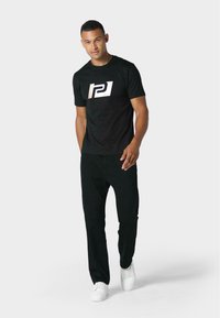 Camiseta negra con un logo blanco, combinada con pantalones negros y zapatillas blancas. La tela parece suave y el ajuste es informal.