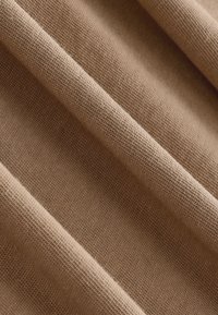 Primo piano di tessuto beige con trama visibile e pieghe morbide che creano linee diagonali e ombre.