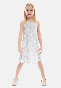 Weißes Kleid mit kleinem blauen Blumenmuster, A-Linie, Spaghetti-Träger. Das Model trägt beige Schuhe mit Riemen.