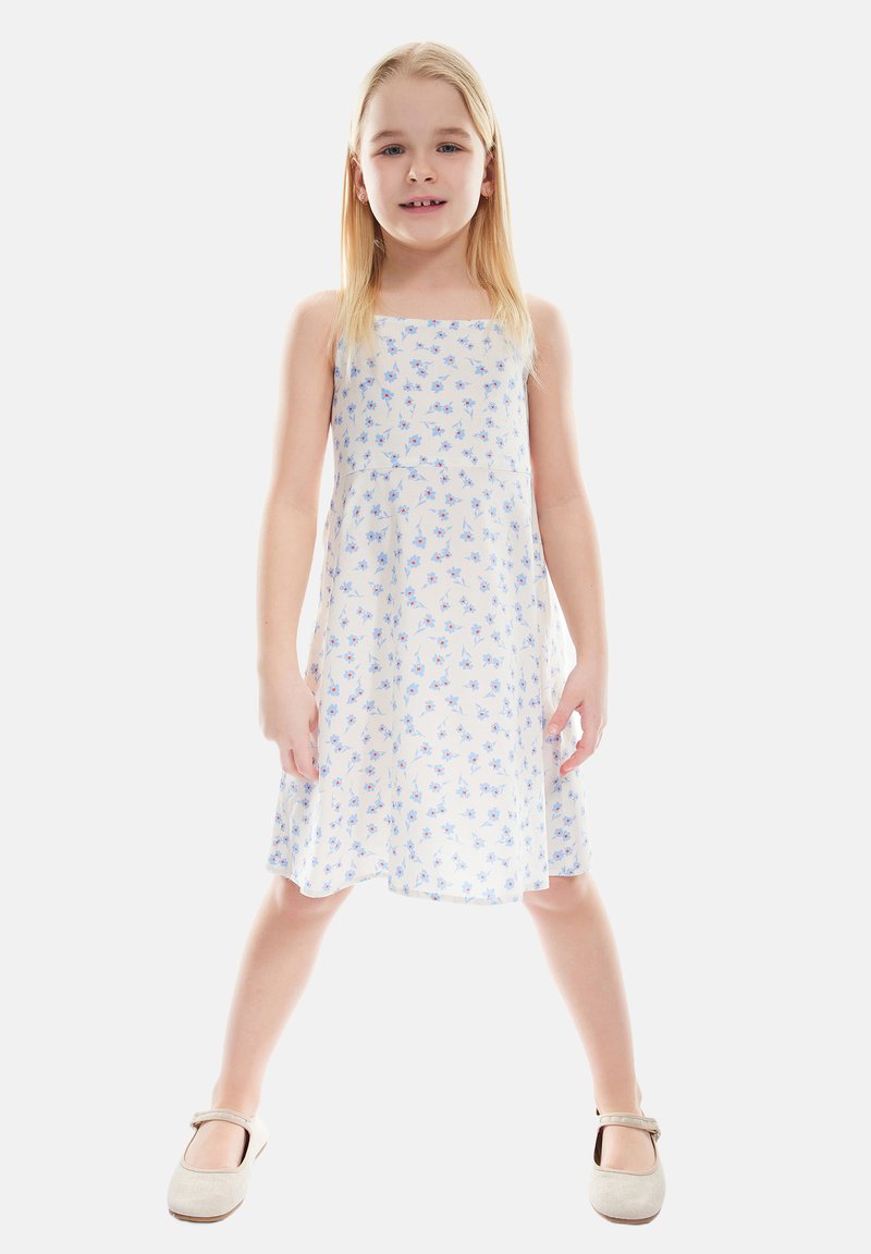 Weißes Kleid mit kleinem blauen Blumenmuster, A-Linie, Spaghetti-Träger. Das Model trägt beige Schuhe mit Riemen.