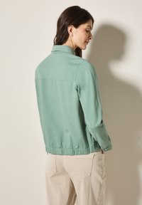 Lichtgroene jas met een kraag, elastische zoom en lange mouwen. Gemaakt van zachte stof. Model draagt lichtbeige broeken.