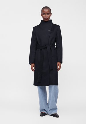 Personne portant un long manteau noir ceinturé avec un col haut, un jean large bleu clair et des chaussures noires pointues, debout devant un fond uni.
