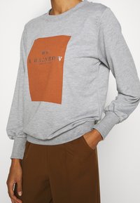 Grå sweatshirt med långa ärmar som har en brun rektangulär grafik och texten "VAR EN REGNBÅGE". Mjuk bomullsmaterial med ribbade muddar.