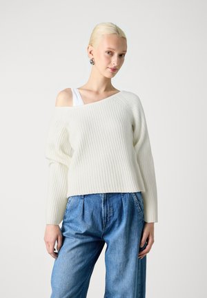 Light cream ribbed sweater with an off-shoulder design, paired with loose-fitting blue denim pants. Soft, textured fabric with long sleeves.