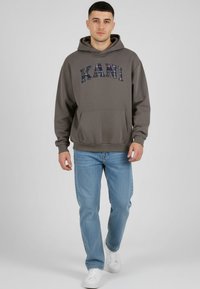 Noor mees kõnnib, kandes halli KANI dressipluusi, helesiniseid pükse ja valgeid tossusid, taust on hele ja lihtne.