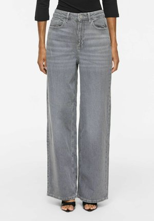 Person iført grå jeans med høj talje og vide ben, kombineret med en sort top med lange ærmer og sorte åbne hæle mod en hvid baggrund.