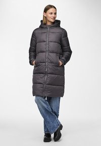 Grå pufferjakke med hette, glidelås foran og to lommer. Har et quiltet design og lengre lengde. Kombinert med løse denimjeans.