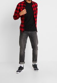 Czerwona i czarna flanelowa koszula w kratę nałożona na czarny T-shirt, w połączeniu ze szarymi dżinsami i czarnymi trampkami, podwinięte mankiety przy kostkach.