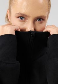 Mustast pehmest materjalist lukuga kapuutsiga pusa, millel on soonilised varrukaotsad ja kõrge krae. Suur plaan inimese näost, mis on osaliselt riidevarjuga varjatud.