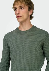 Pull vert tricoté texturé avec un col rond et des manches longues, présentant un motif en grille serrée pour un intérêt visuel accru.