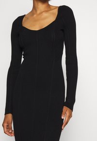 Schwarzes, geripptes Langarmkleid mit quadratischem Ausschnitt, das vertikale Strickmuster und eine enganliegende Silhouette aufweist. Weiche Textur sichtbar.