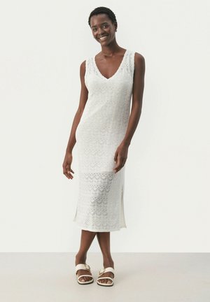 Weißes gestricktes Kleid mit V-Ausschnitt, das mit einem strukturierten Muster und Seitenschlitzen versehen ist. Das Modell trägt braune Sandalen mit einer goldenen Schnalle.