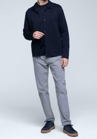 Granatowa pleciona kurtka na guziki z kołnierzem i dwoma przednimi kieszeniami, zestawiona z jasnoszarymi spodniami i ciemnymi trampkami z gumową podeszwą.