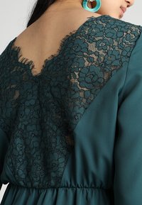 Tiefblaues Kleid mit einem Spitzenrücken mit Blumenmustern, einem V-Ausschnitt und geraffter Taille. Glatter Stoff mit strukturierten Spitzenakzenten.