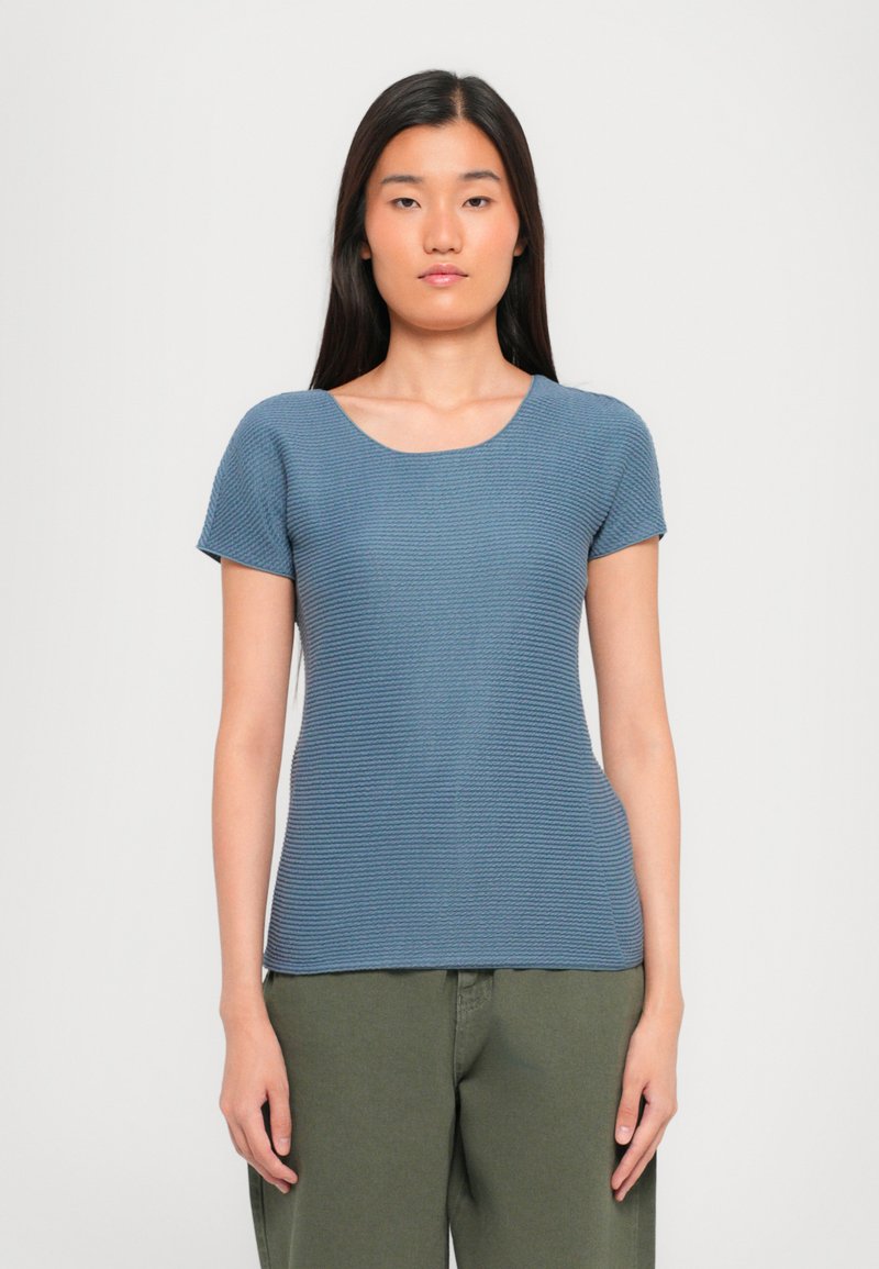 Short-sleeve, round-neck top in textured blue fabric, fitted silhouette, with a slightly ribbed pattern. Paired with olive green pants.