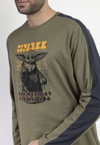 Camisa de manga larga verde oliva con un acento de manga azul marino. Presenta una impresión gráfica de un personaje con el texto "Pequeño pero Poderoso".