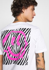 White t-shirt featuring a large pink smiley face graphic against a black and white zebra stripe background. Short sleeves, round neck.