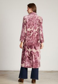 Manteau floral long en rose pâle, avec des motifs bordeaux profonds. Fabriqué en tissu texturé avec un col cranté et des manches longues. Porté sur un jean noir.