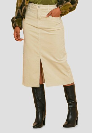 Jupe en denim beige avec une fente à l'avant, deux poches et une fermeture à bouton. Associée à des bottes noires à talons hautes jusqu'aux genoux.