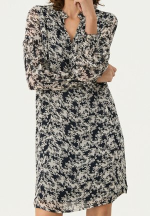 Kobieta w sukience sięgającej kolan z długimi rękawami, w czarno-biały kwiatowy wzór, stojąca z założonymi ramionami i ręką opartą na brodzie.