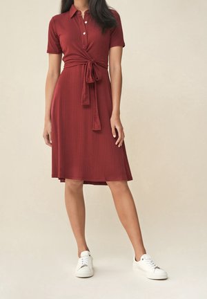 Femme portant une robe courte bordeaux à manches courtes et longueur genoux avec un lien à l'avant, associée à des baskets blanches sur un fond uni.