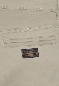 Pantalones de tela beige con un bolsillo trasero que presenta una etiqueta "SIGNAL" en azul y dorado, acentuada por costuras en contraste.