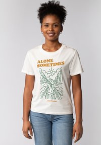 Weißes Baumwoll-T-Shirt mit grünem Linienstil, das eine Figur vor einem bergigen Hintergrund zeigt, zusammen mit orangefarbener Schrift, die "MANCHMAL ALLEIN... aber das ist okay." liest.
